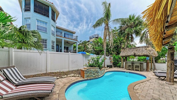 A swimming pool with lounge chairs, a thatched-roof structure, and a white fence. - Tuscan Tides - Custom Pool Home - powered by Island Real Estate (Holmes Beach)