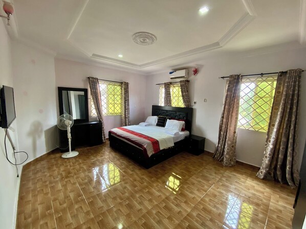 A bedroom with a large bed, a flat-screen TV, a standing fan, and a window with curtains. - 6 Bedroom Serviced Apartments In House in Benin (Benin City)