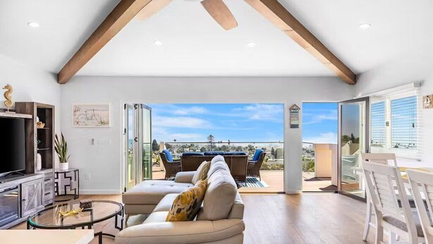 Un salón amplio con techo alto, un sofá grande, una mesa de centro de vidrio y una televisión. Hay un balcón con mobiliario exterior y vistas del paisaje.