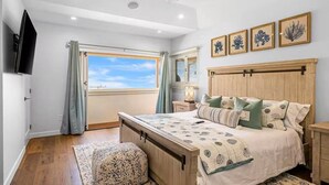 Un dormitorio con cama, ventilador de techo, ventana con cortinas, mesita de noche y cuadros en la pared.