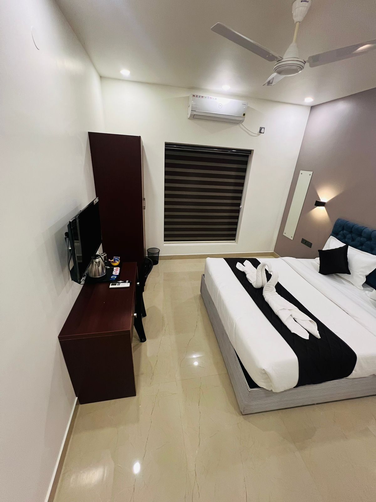 A modern hotel room with a bed, a desk, a wardrobe, a window with blinds, a ceiling fan, and an air conditioning unit.