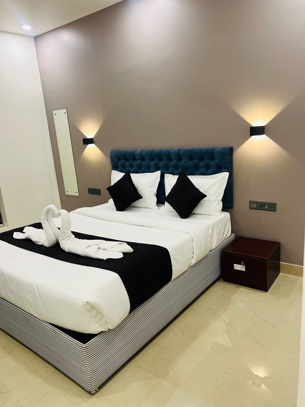 A modern hotel room with a large bed, black and white bedding, a blue tufted headboard, and two wall-mounted light fixtures.