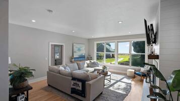 A spacious living room with a large window, a sofa, a coffee table, and a TV mounted on the wall.