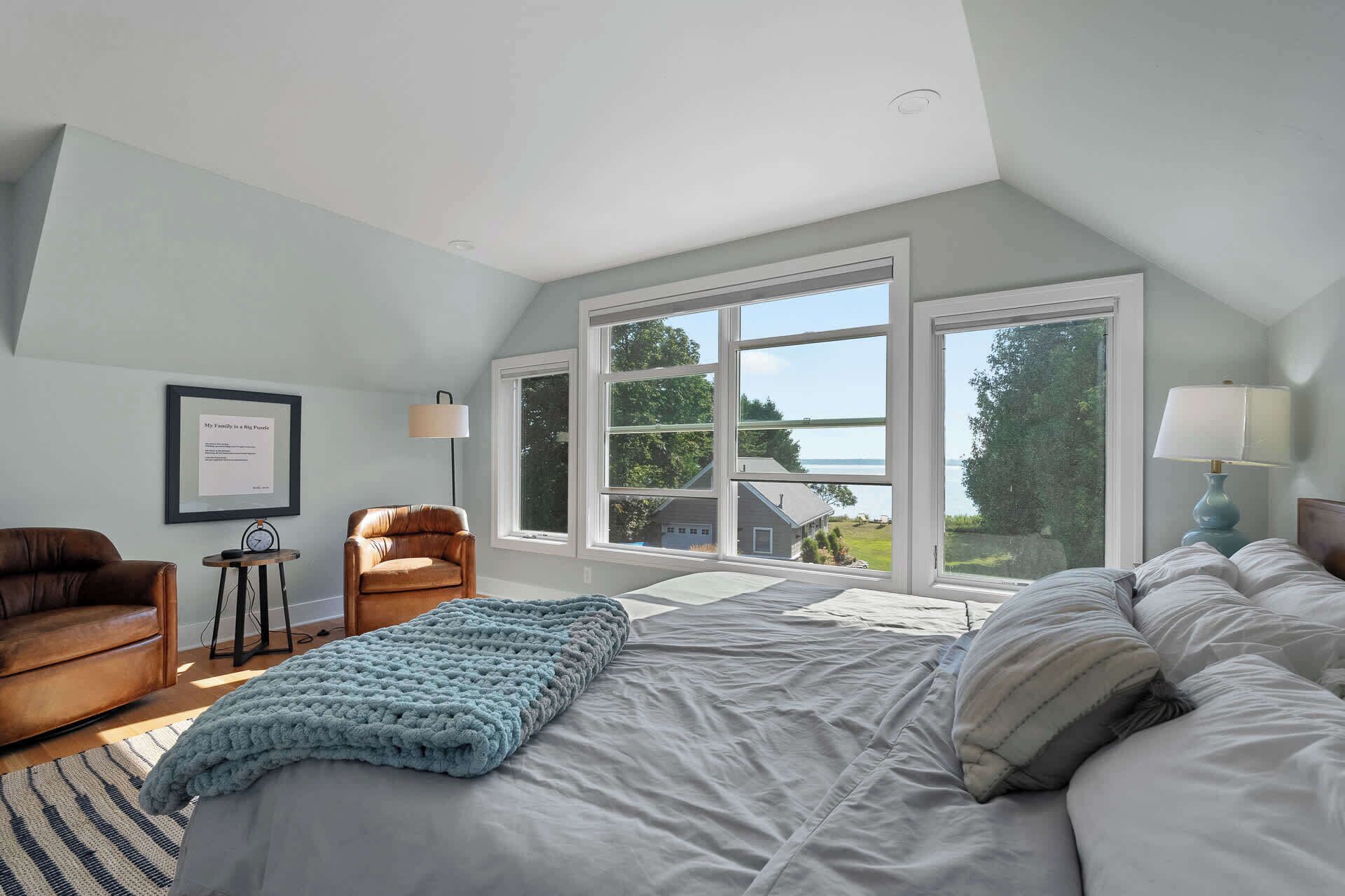 A bedroom with a large bed, a chair, a desk, and a window with a view of a house and trees.
