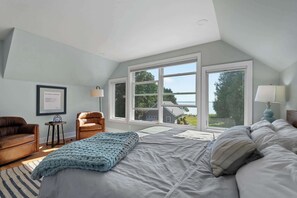 A bedroom with a large bed, a chair, a desk, and a window with a view of a house and trees. - Downtown Baileys Harbor Waterfront: Limestone Edge (Baileys Harbor)