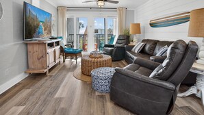 A living room with a TV, leather sofa, wicker ottoman, and a ceiling fan.