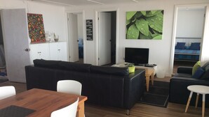 Un salon moderne avec un canapé noir, des chaises blanches, une table à manger en bois et un téléviseur posé sur un meuble en bois.