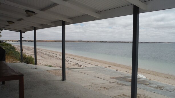 Un espace extérieur abrité, doté d’un sol en béton, de poteaux métalliques et offrant une vue sur une plage et l’eau.