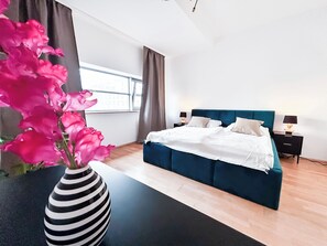 A modern bedroom with a large bed, bedside tables, a vase of flowers, and a view of the city through the window.