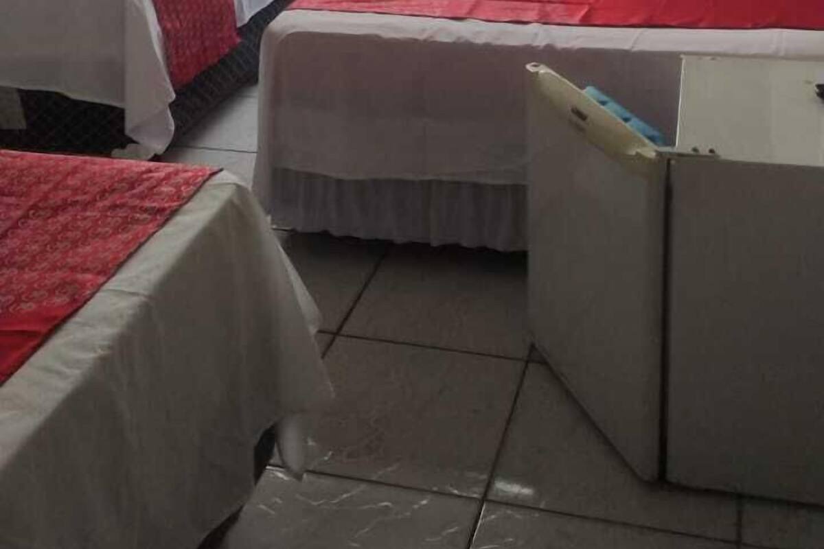 Habitación de hotel con dos camas, cada una con una almohada roja y una colcha blanca. Hay un pequeño armario blanco en la esquina y una ventana con cortinas.