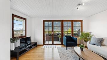 Ett modernt vardagsrum med en platt-TV, en soffa, en fåtölj och ett soffbord. Det finns stora skjutdörrar i glas som leder ut till en balkong med utsikt över bergen.