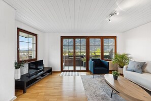 Sala de estar moderna com televisão de tela plana, sofá, poltrona e mesa de centro. Há grandes portas de vidro deslizantes que levam a uma varanda externa com vista para montanhas.