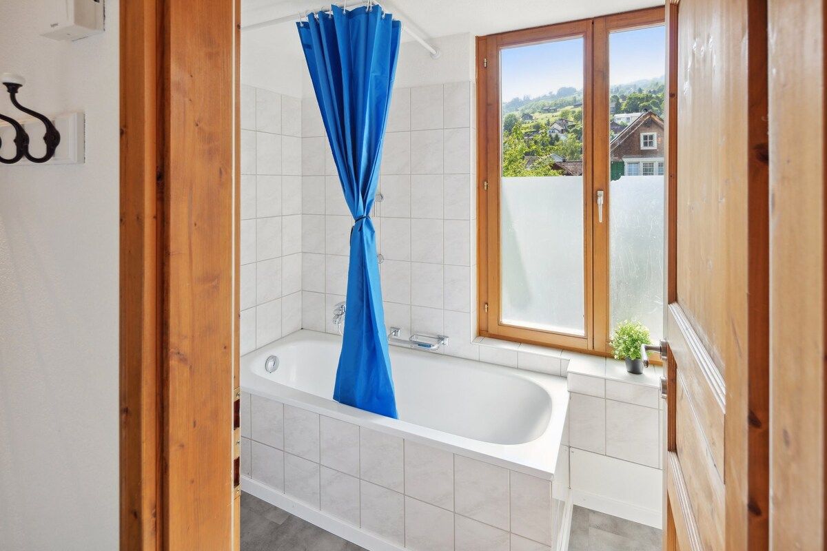A bathroom with a bathtub, a shower curtain, and a window with a view of houses.