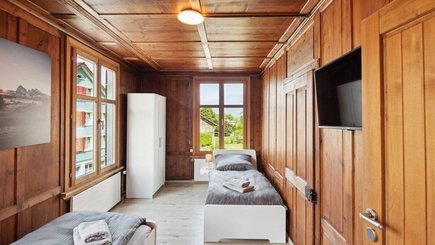 A wooden cabin room with two beds, a TV, and a window with a view of greenery.