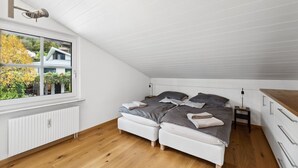 Une chambre avec un plafond en pente, une grande fenêtre donnant sur des maisons et des arbres, deux lits avec une literie grise, un radiateur blanc et un plancher en bois.