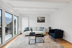 A modern living room with a white sofa, two black coffee tables, a blue armchair, and a large window with a view of mountains. - FENUS View over Liechtenstein | Parking | Kitchen (Buchs)