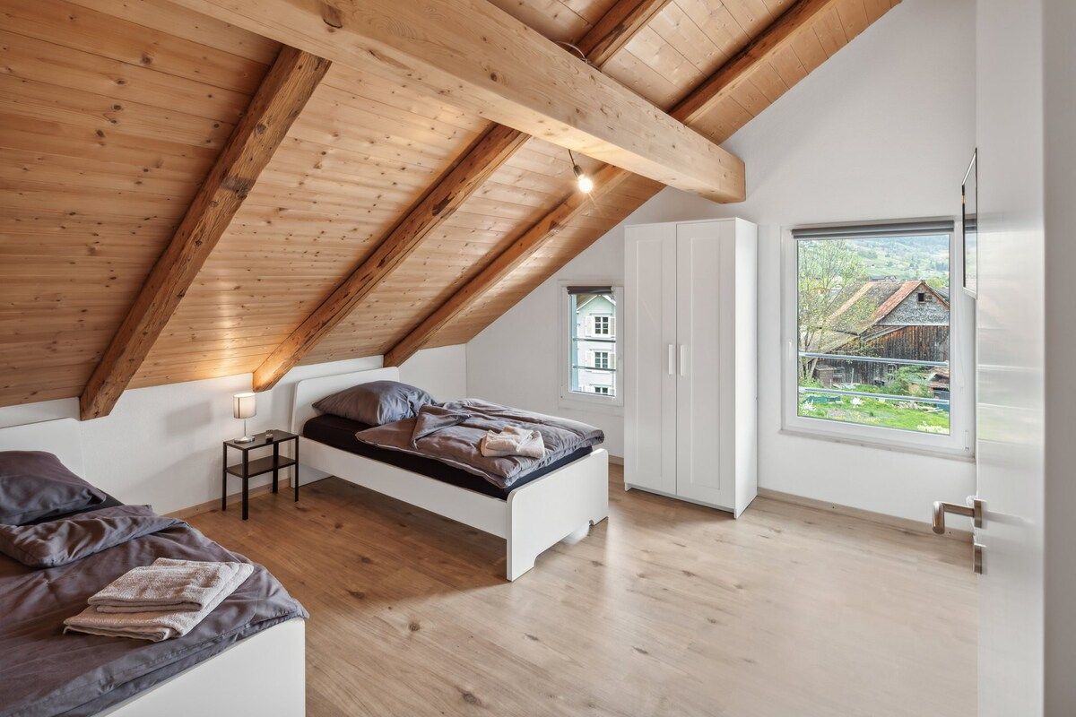 Une chambre avec un plafond en pente, un lit, une table de chevet, une fenêtre donnant sur des maisons et une armoire.