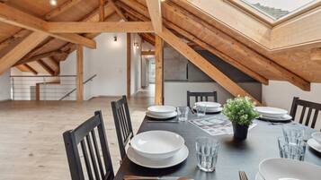 Une salle à manger avec un plafond haut et des poutres en bois, une longue table dressée pour quatre personnes et une lucarne au-dessus.