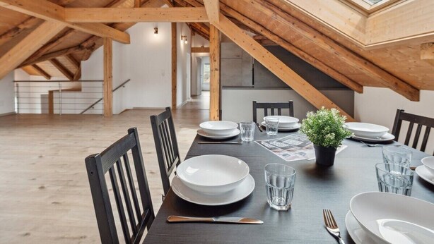 Un comedor con techo alto y vigas de madera, una mesa larga dispuesta para cuatro personas y un tragaluz arriba.