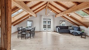 Un loft amplio con techo alto y vigas de madera expuestas, una zona de comedor con mesa y sillas, y un sofá de color oscuro en la sala.