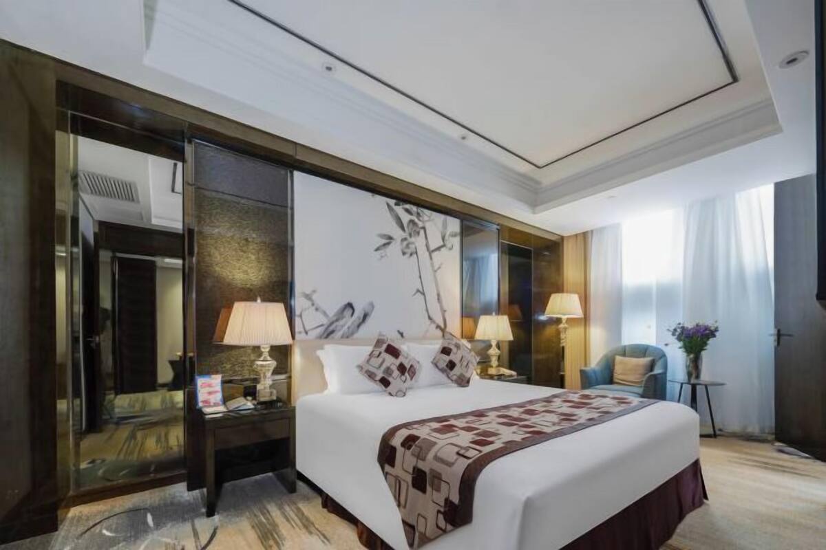 A hotel room with a large bed, two bedside tables with lamps, a chair, a glass partition, and a large window with curtains.