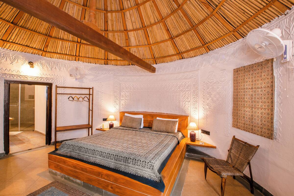 Quarto com cama de madeira, roupa de cama estampada, teto de palha e painéis decorativos na parede.
