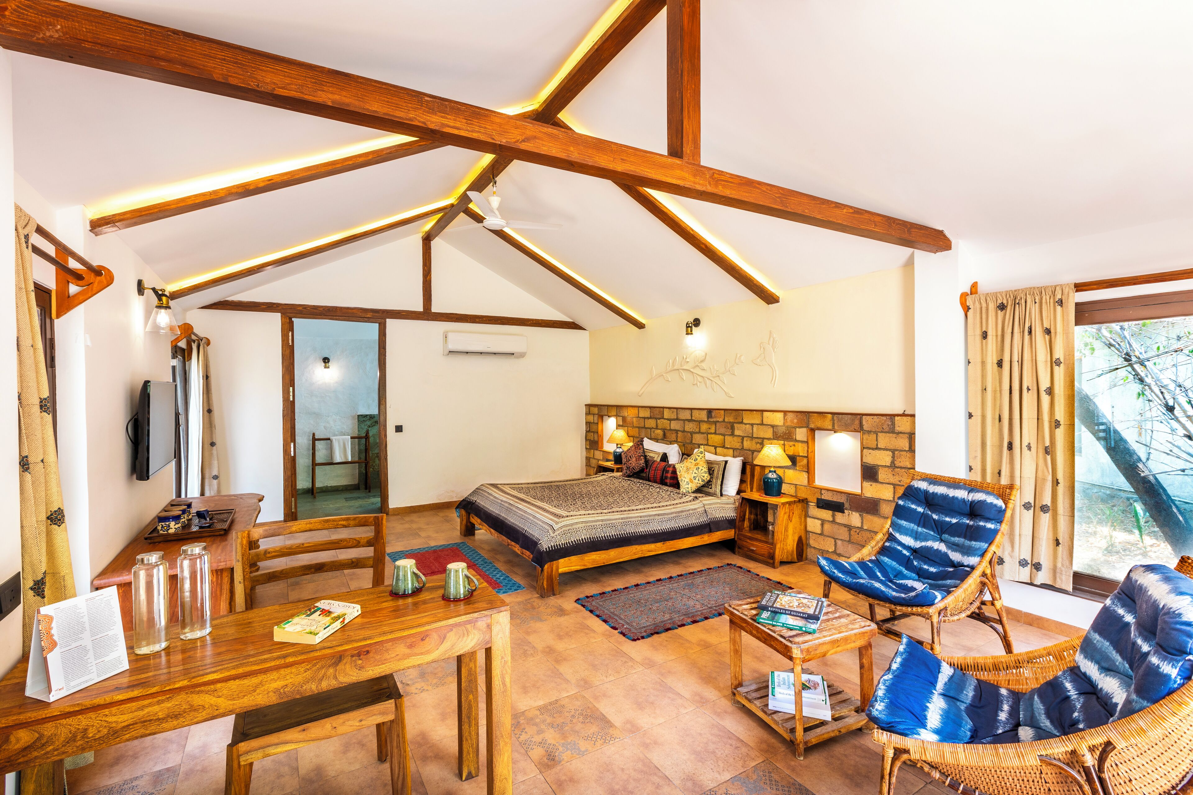 Une pièce spacieuse avec un plafond haut, des poutres en bois, un lit avec une tête de lit, deux fauteuils en osier, une petite table et une fenêtre avec des rideaux.