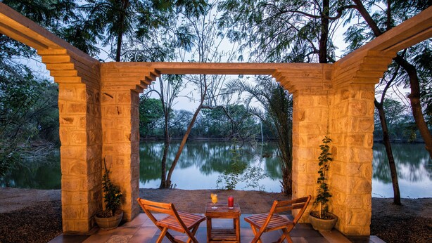 Une pergola en bois avec une table et des chaises, donnant sur un lac, et entourée de murs en pierre et de plantes en pot.