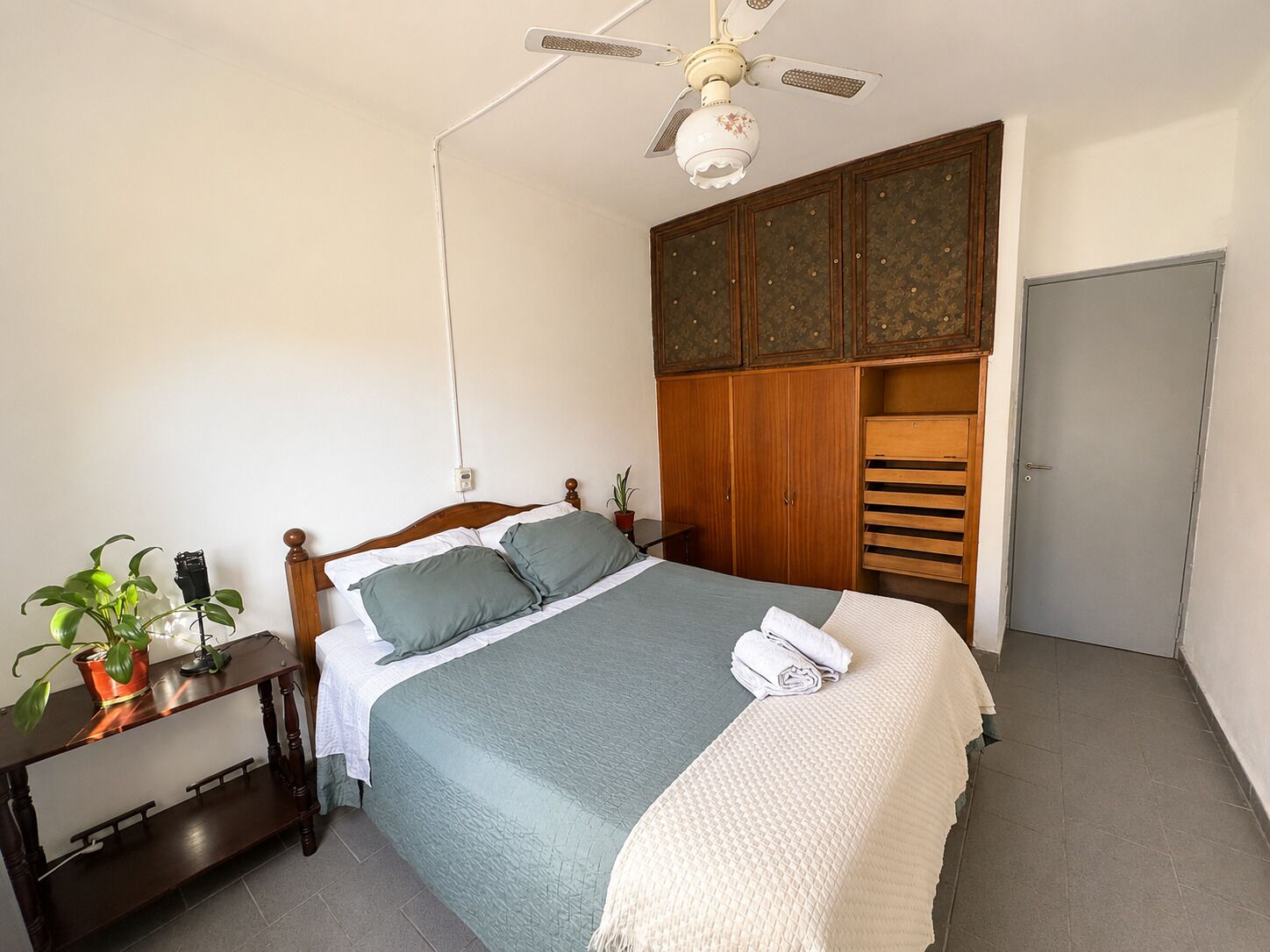 Un dormitorio con cama, mesita de noche con una planta, armario y ventilador de techo.