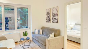 A modern living room with a grey sofa, a small round table with a plant, and a bed in the adjacent room.