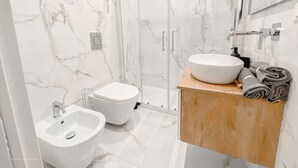 A modern bathroom with a marble wall, a glass shower enclosure, a white sink, and a bidet.