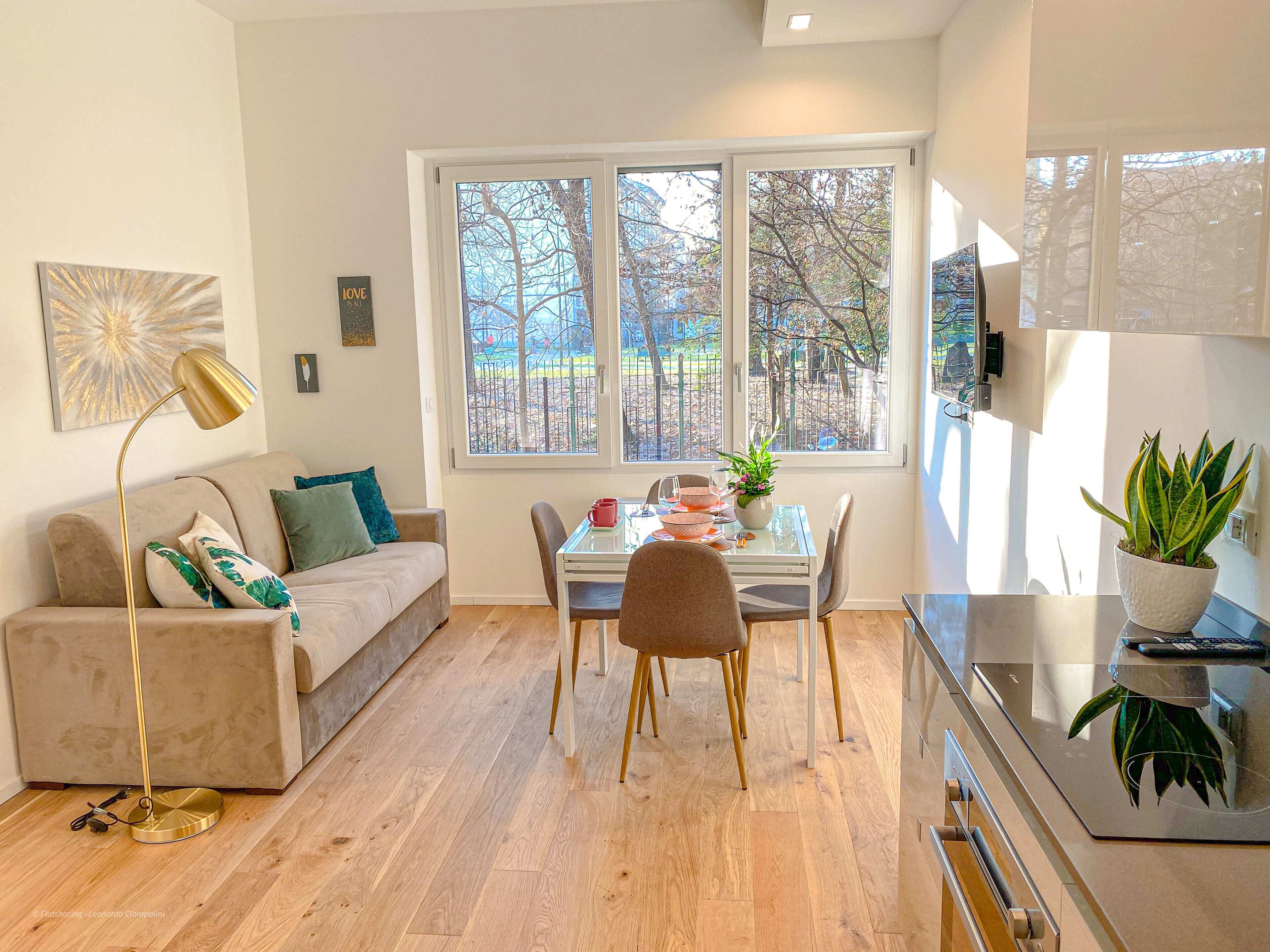 A modern living room with a sofa, a dining table, chairs, a floor lamp, and a view of trees through large windows.