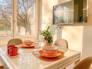 Une table à manger avec des bols et des tasses ornés de motifs orange, un vase rempli de fleurs et un téléviseur fixé au mur.