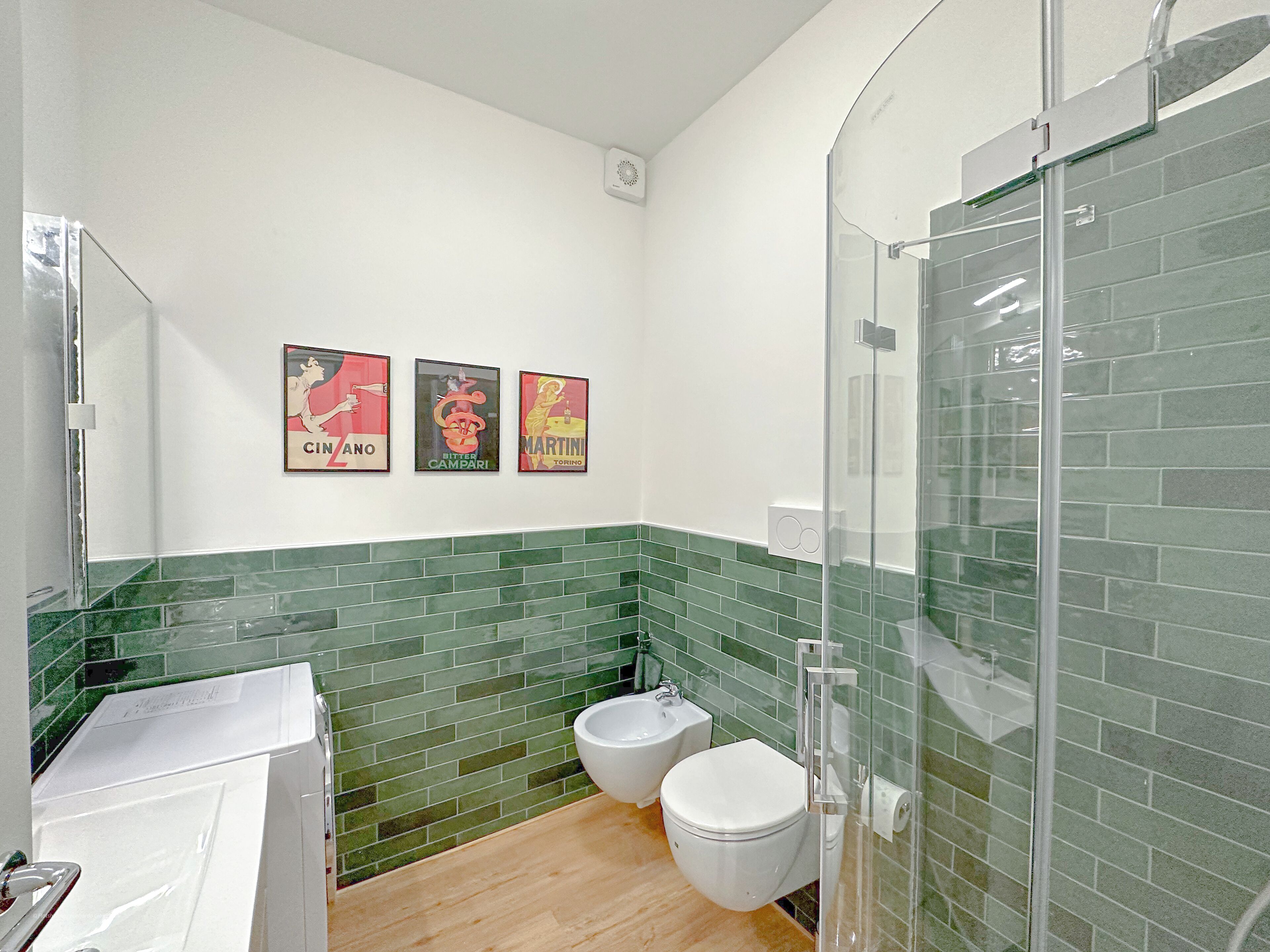 Ein Badezimmer mit Toilette, Bidet und einer Duschkabine aus Glas. An der Wand hängen drei Vintage-Poster.