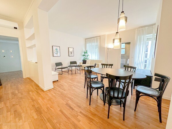 Un vaste espace de vie doté d’un plancher en bois, d’une table à manger avec des chaises, d’un canapé et d’une grande fenêtre agrémentée de rideaux.