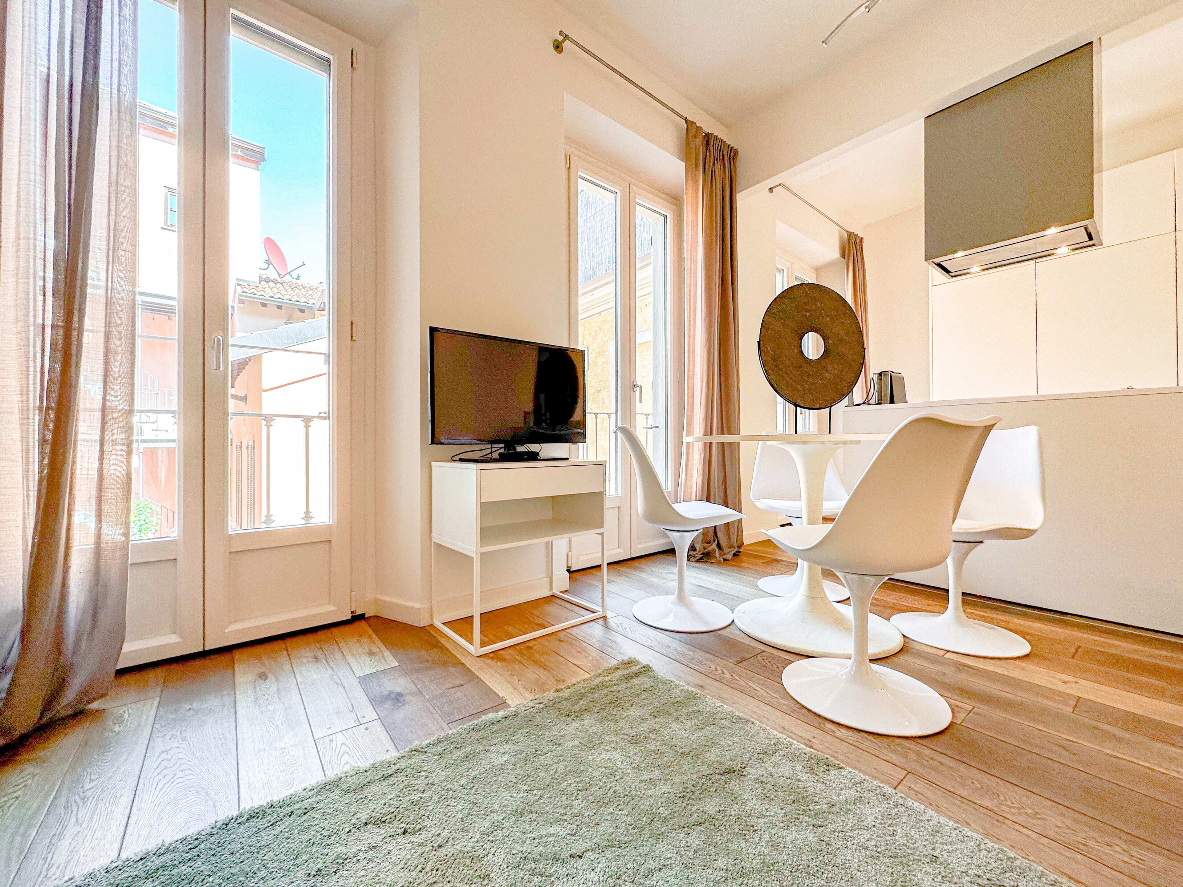 A modern living room with a TV, white chairs, a round table, and a large window with curtains.