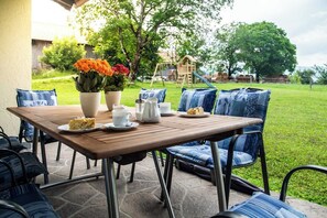Een terras buiten met een houten tafel gedekt voor het ontbijt, omringd door blauwe fauteuils.