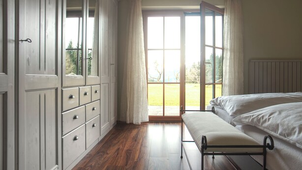 Un dormitorio con un ventanal grande, suelo de madera, una cama, un banco y un armario.
