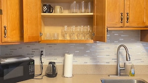 A kitchen with wooden cabinets, a microwave, a kettle, a paper towel holder, a sink, and a dish soap bottle.