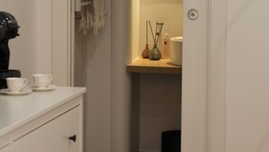 Een smalle gang met een witte linnenkast, een spiegel en een klein plankje met een vaas en een fles.