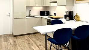 Een moderne keuken met witte kastjes, een ingebouwde oven, een magnetron en een koffiezetapparaat. Er staan twee blauwe stoelen aan een witte tafel.