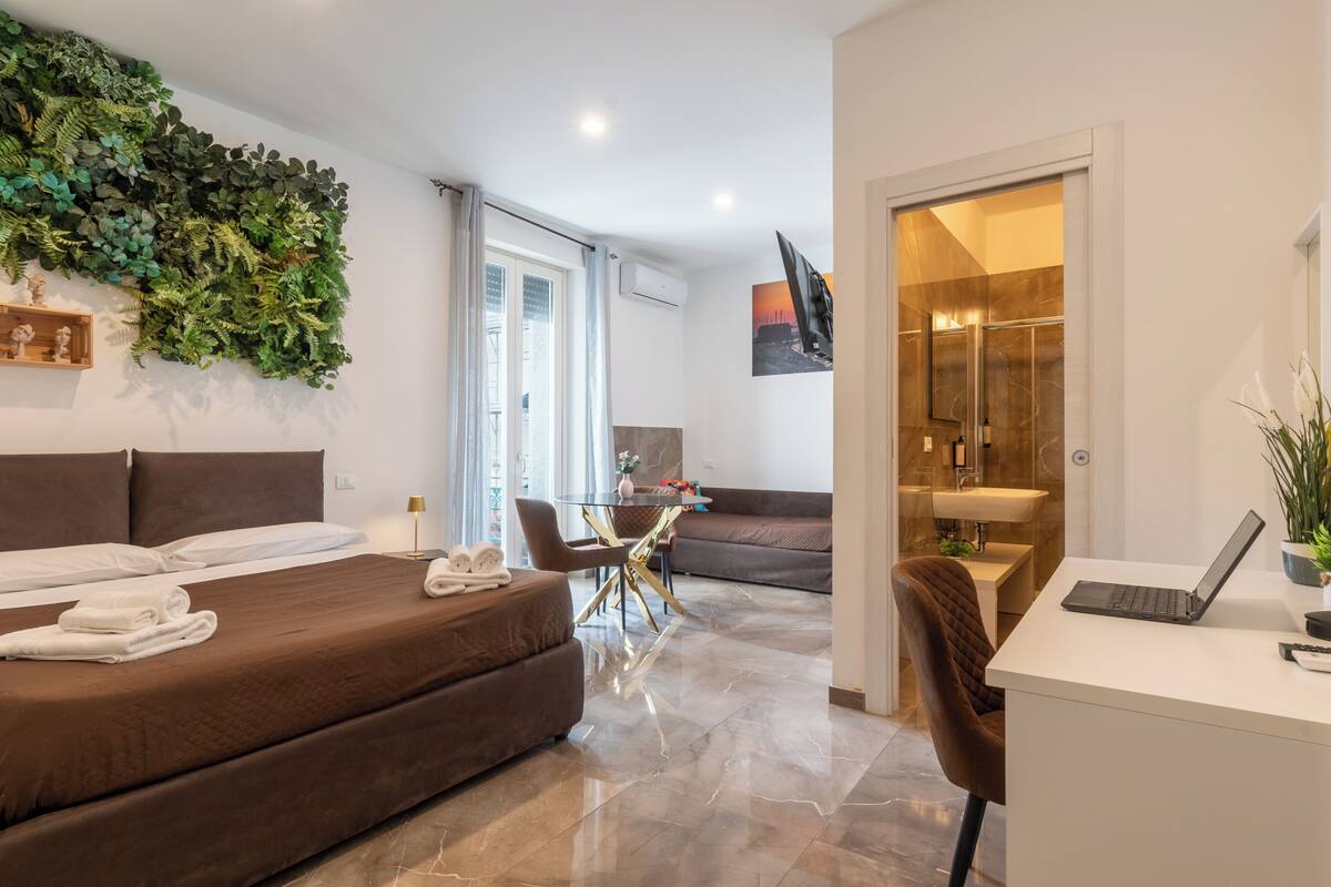A modern hotel room with a bed, a desk, a seating area, and a bathroom.