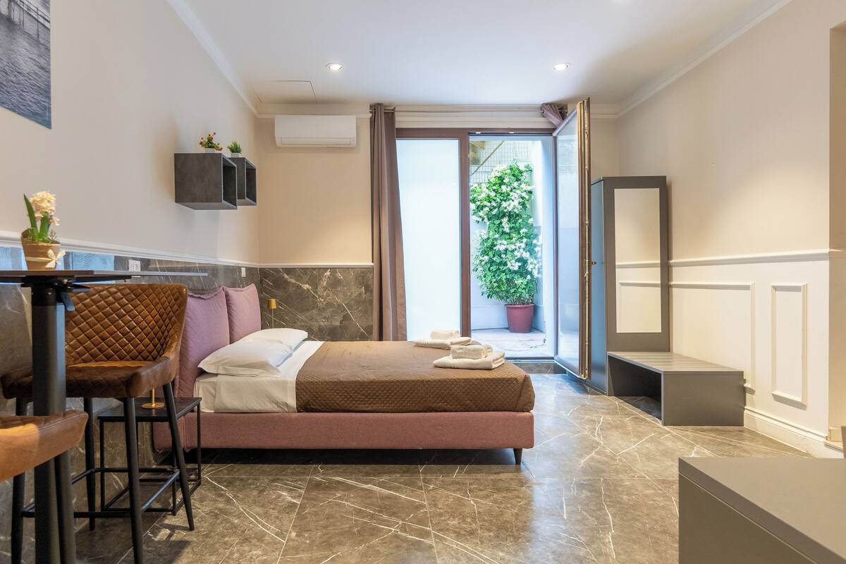 A modern hotel room with a bed, a seating area with a chair and table, a potted plant, and a large mirror.