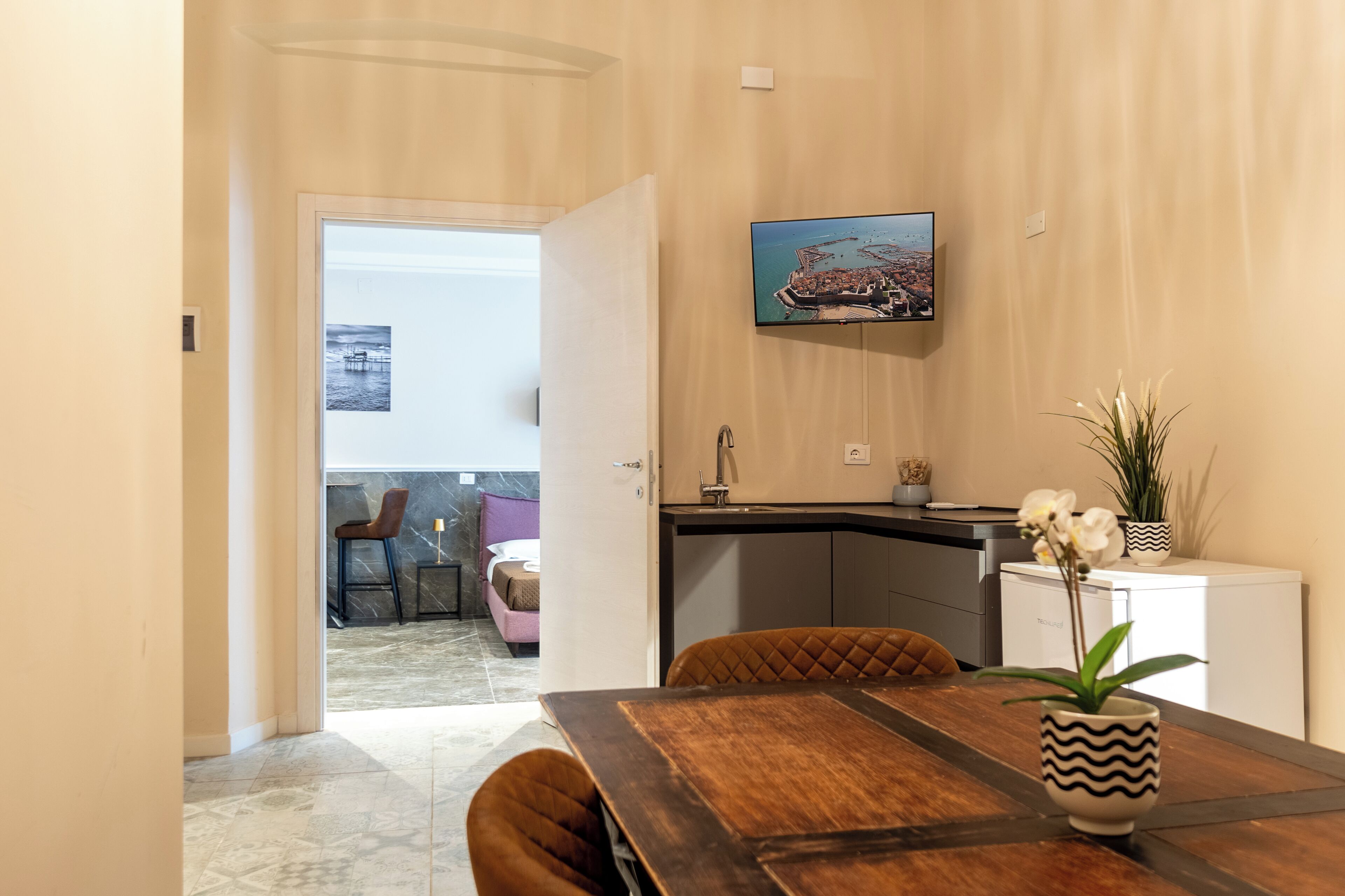 A dining area with a wooden table, chairs, a vase with flowers, a television mounted on the wall, and a view of a room with a bed and a chair.