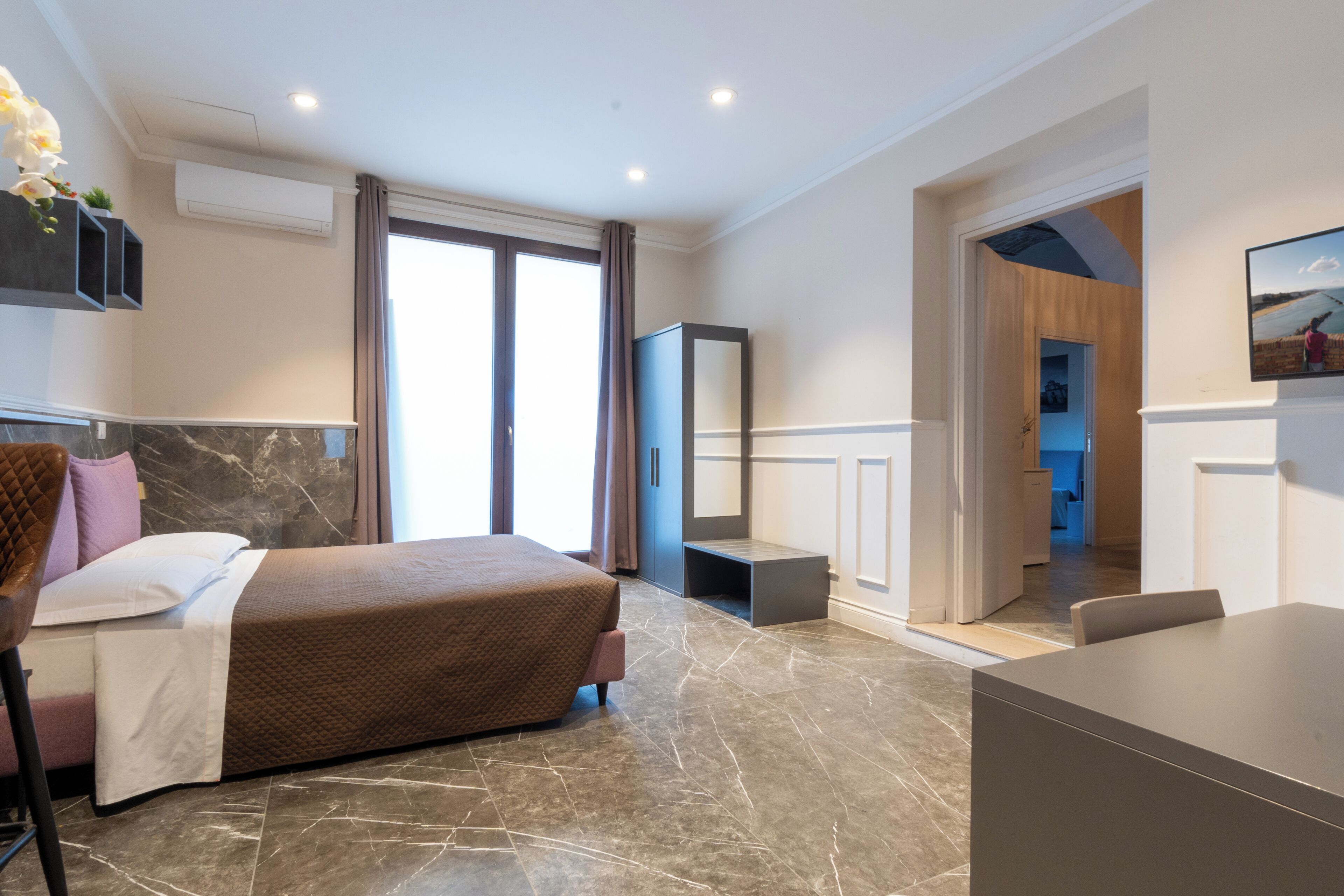 Une chambre d’hôtel moderne équipée d’un lit, d’un bureau, d’une chaise, d’une télévision et d’une salle de bain visible à travers une porte ouverte.