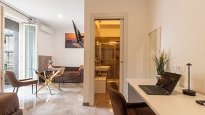 A modern interior with a dining area, a sofa, a laptop on a desk, and a bathroom visible through an open door. - Suebi Central - Deluxe Rooms & Family Suites (Termoli)