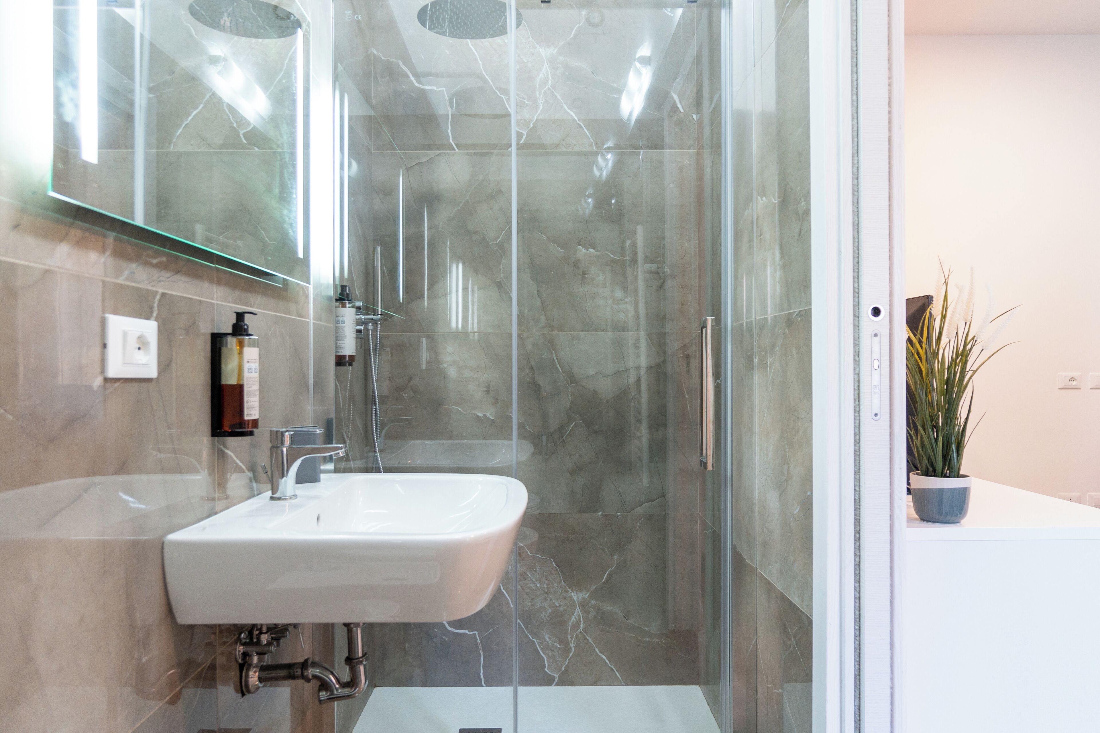 Un baño moderno con cabina de ducha de vidrio, un lavamanos blanco y una planta en maceta.