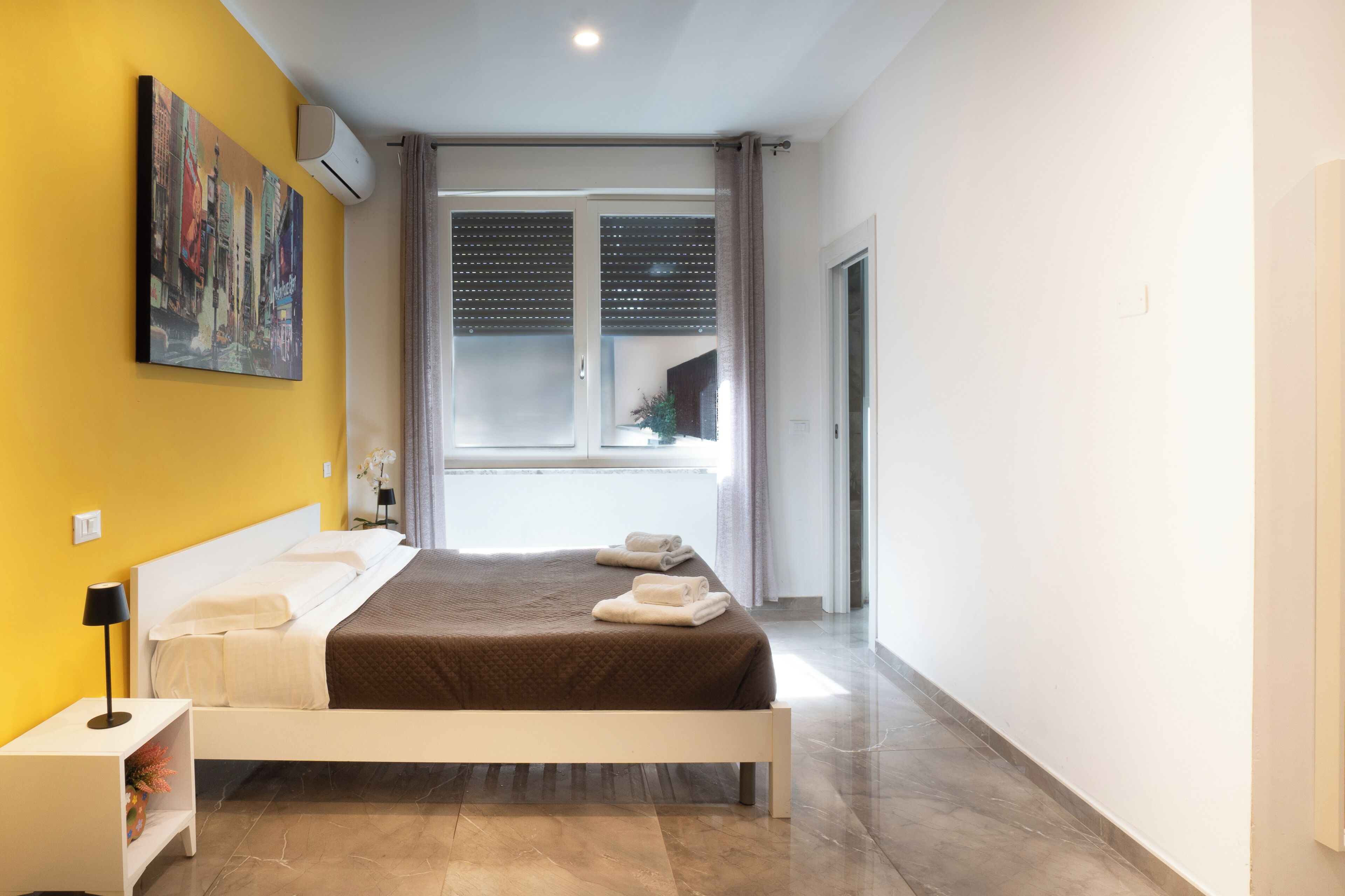 A modern hotel room with a bed, a nightstand, a painting on the wall, and a window with blinds.