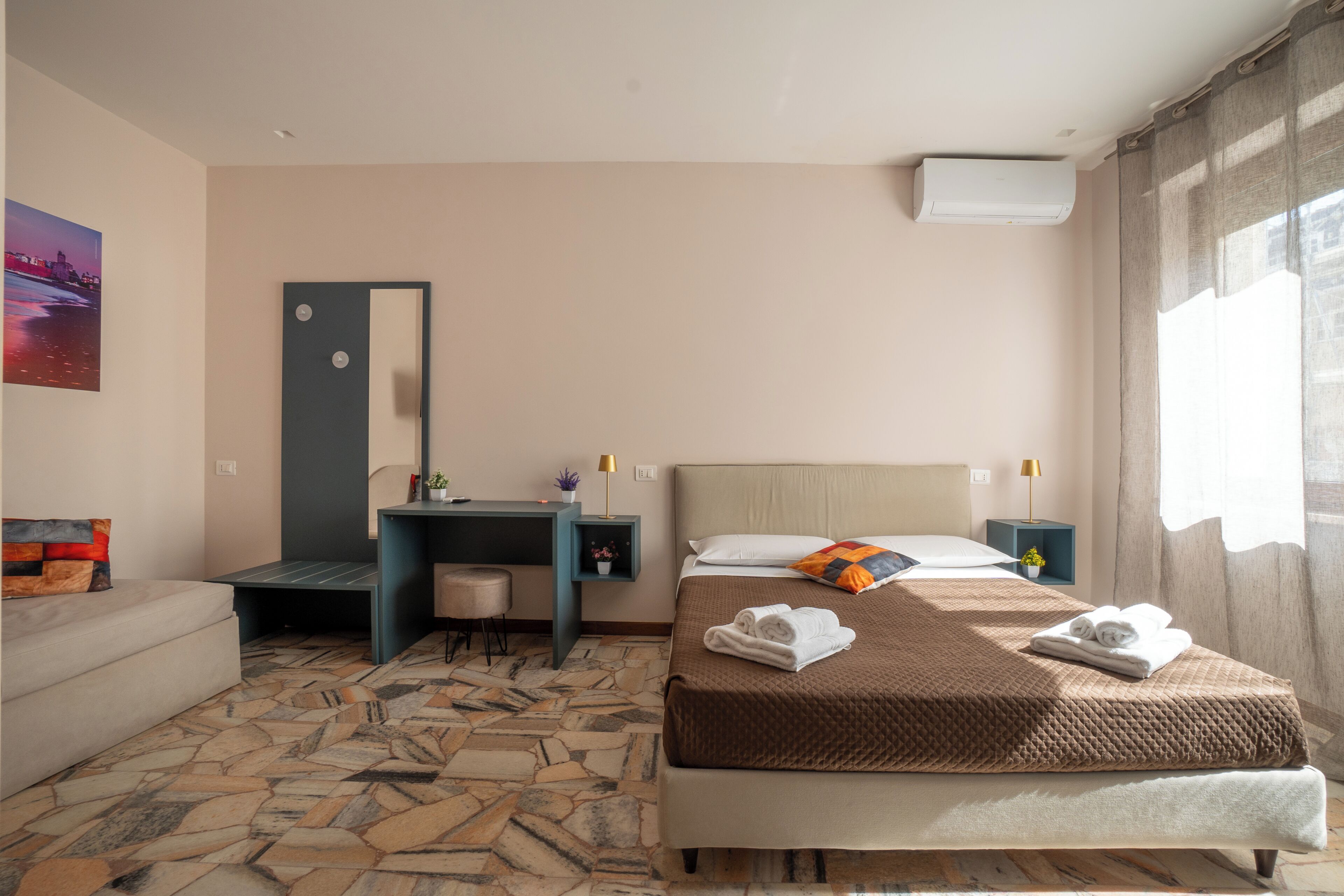 Habitación de hotel con cama, escritorio, silla, mesita, espejo, ventana con cortinas y suelo con diseño de piedra.