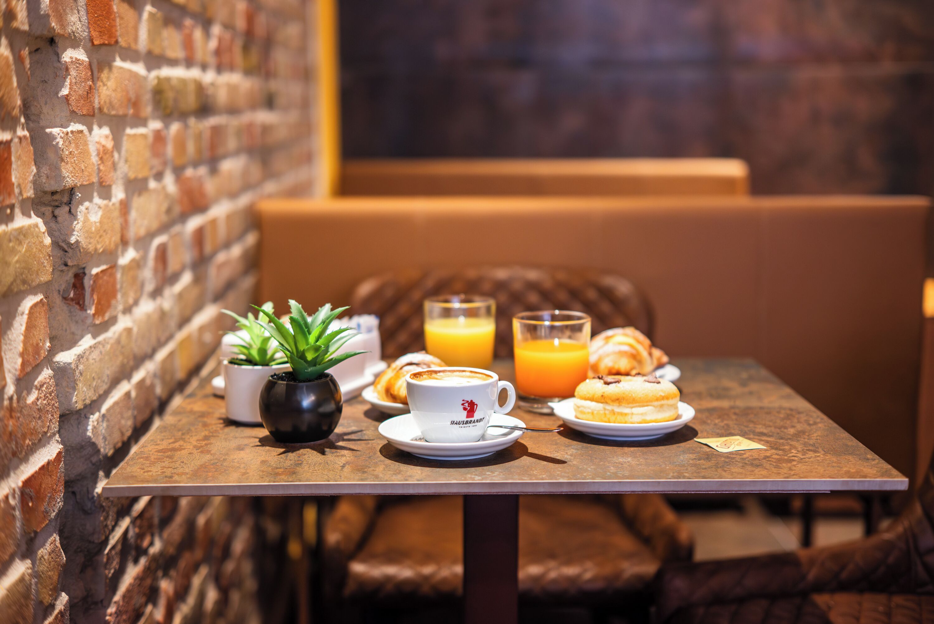Ein Tisch mit Frühstücksleckereien, darunter Croissants, eine Kaffeetasse und ein Glas Orangensaft, vor einer Backsteinmauer.